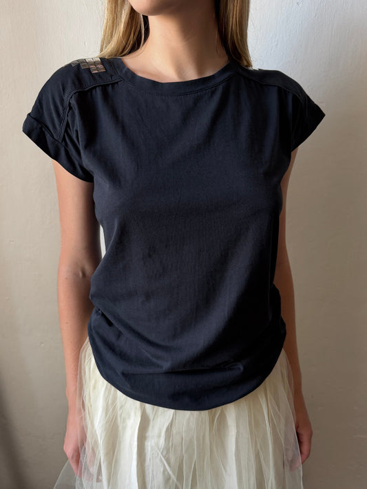 A woman wearing a black vintage short-sleeve t-shirt with structured shoulder panels and a white tulle skirt, standing against a plain beige wall.