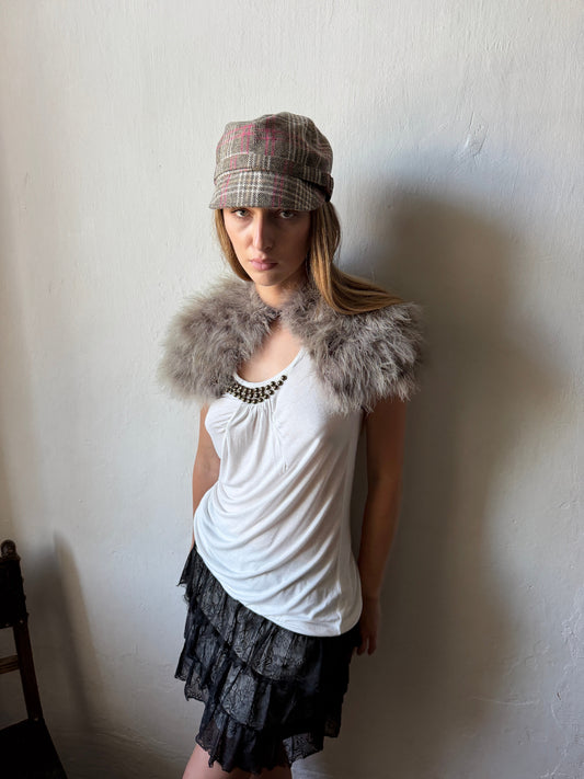 model wearing a white sleeveless top with a fluffy gray faux-fur collar, a black ruffled skirt, and a plaid newsboy cap