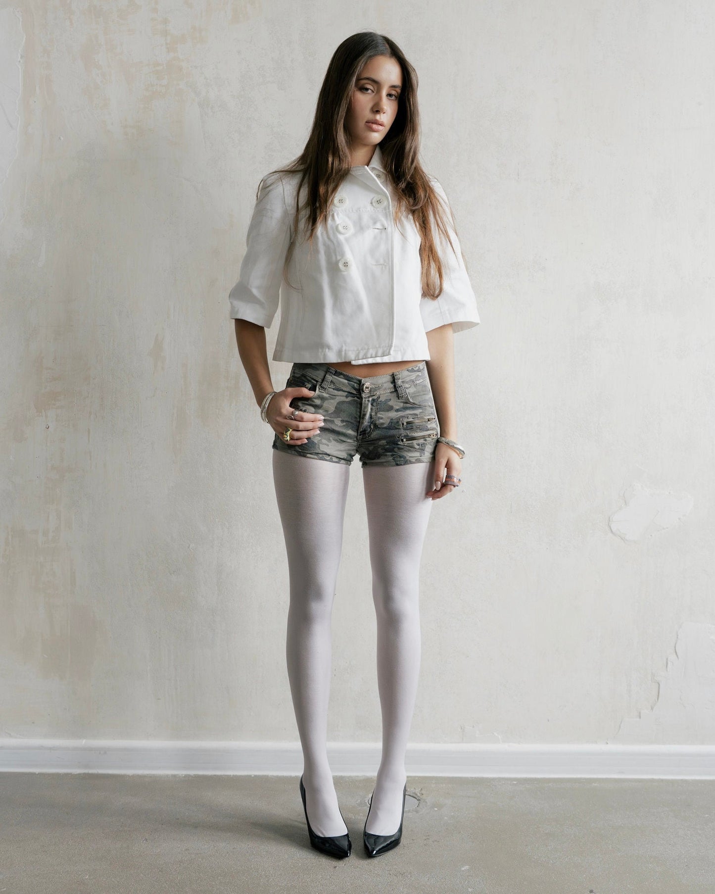Woman wearing a white cropped jacket, gray shorts, and white tights against a plain wall.