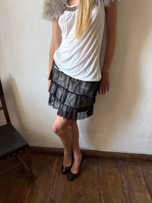 A woman wearing a white sleeveless top with a gray faux-fur collar and a black lace tiered ruffle mini skirt, standing in a room with a wooden floor and off-white wall.