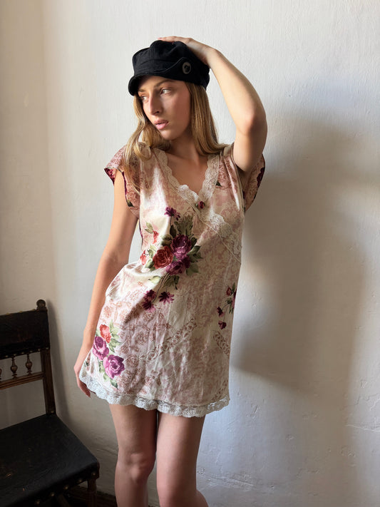 A woman wearing a short-sleeved lace nightgown with a pink and green floral pattern and a black newsboy cap, standing against a white wall.