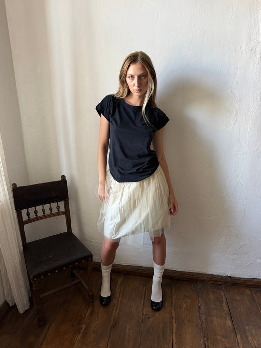 A woman stands in a room with a white wall and wooden floor, wearing a black short-sleeve top, a pleated cream tulle skirt, white socks, and black shoes.