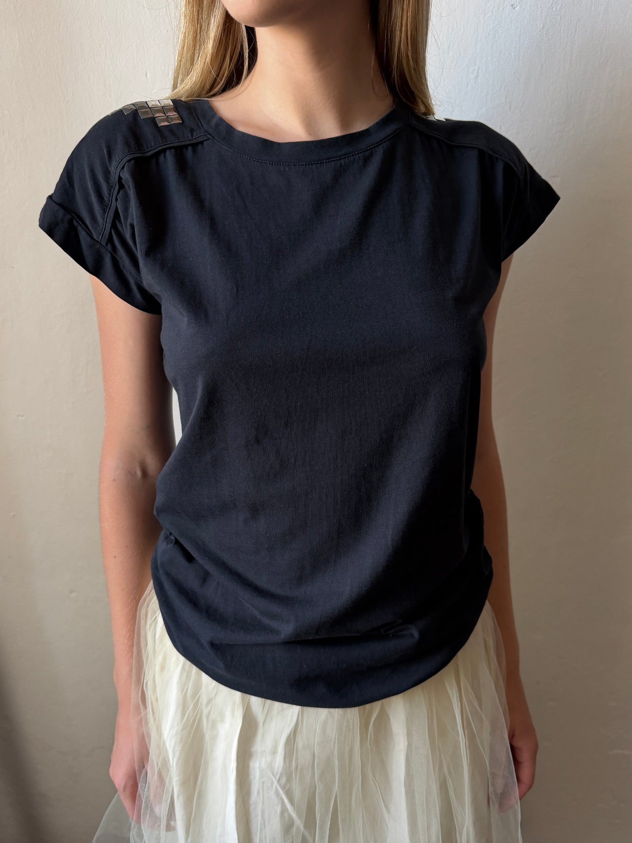 A woman wearing a black vintage short-sleeve t-shirt with structured shoulder panels and a white tulle skirt, standing against a plain beige wall.