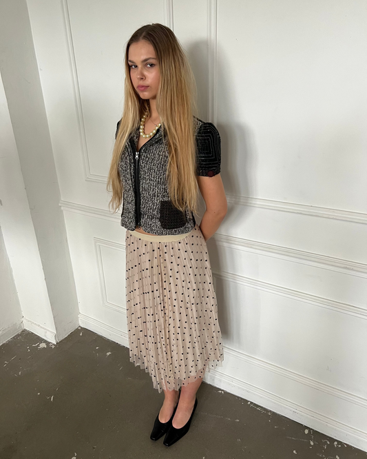 Woman in a stylish outfit with a black top and beige skirt against a white wall.