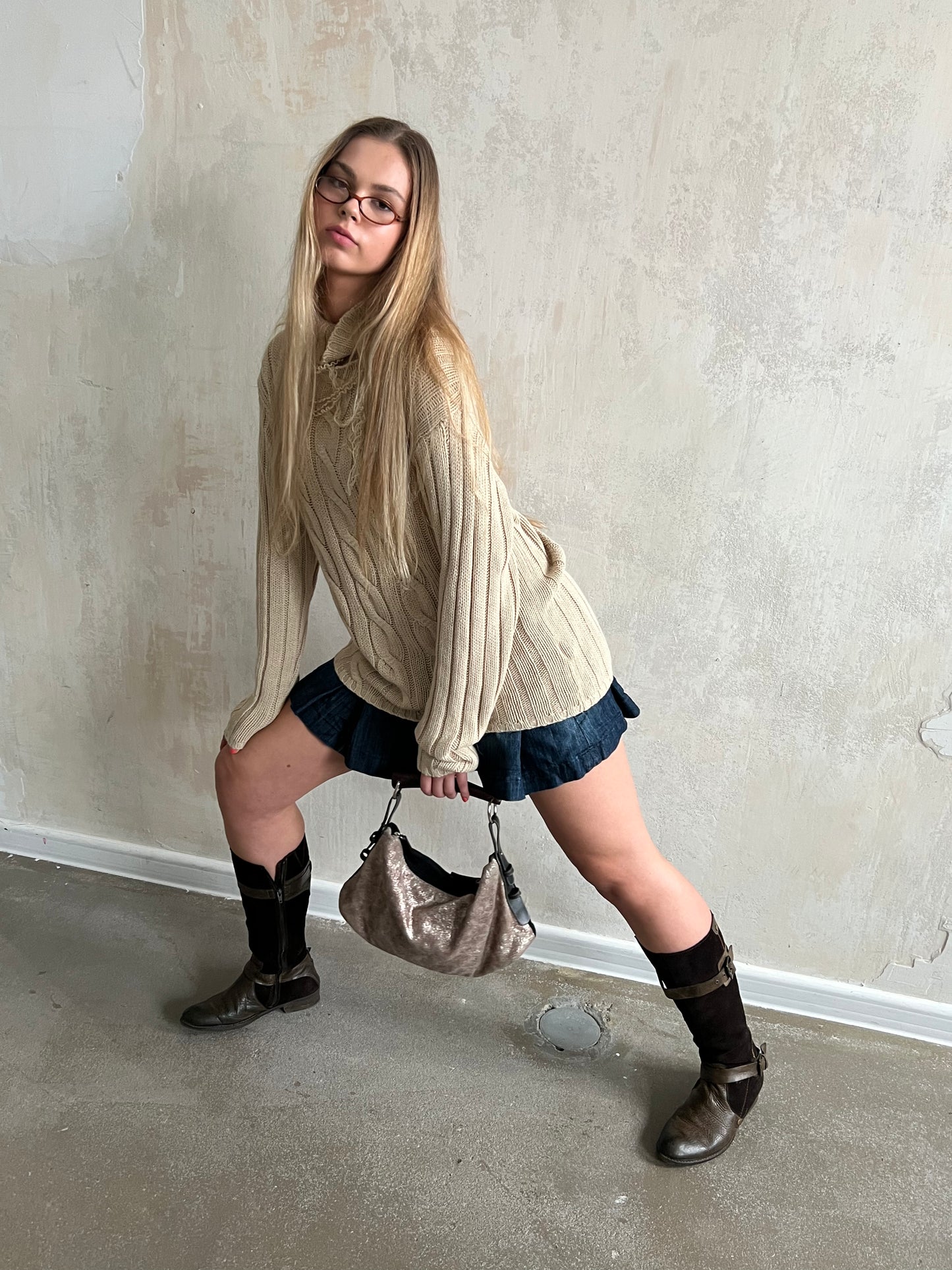 Person wearing a beige sweater, denim skirt, and knee-high boots against a plain wall.