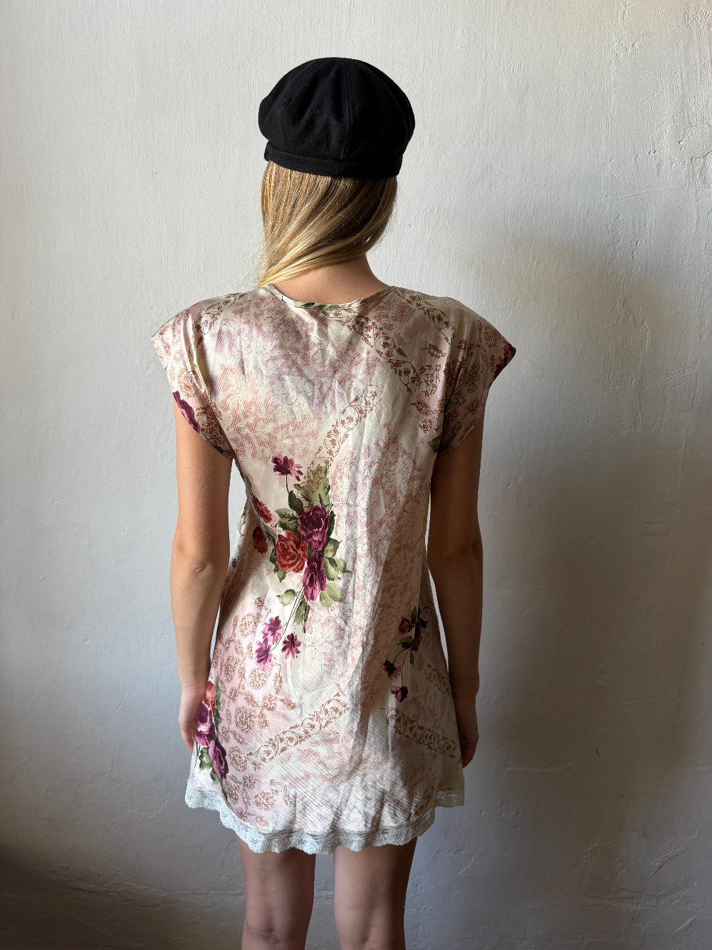 A woman viewed from behind wearing a short-sleeve cream-colored dress with a vintage floral and paisley print in muted red, purple, and green tones, paired with a black beret, standing against a textured off-white wall.