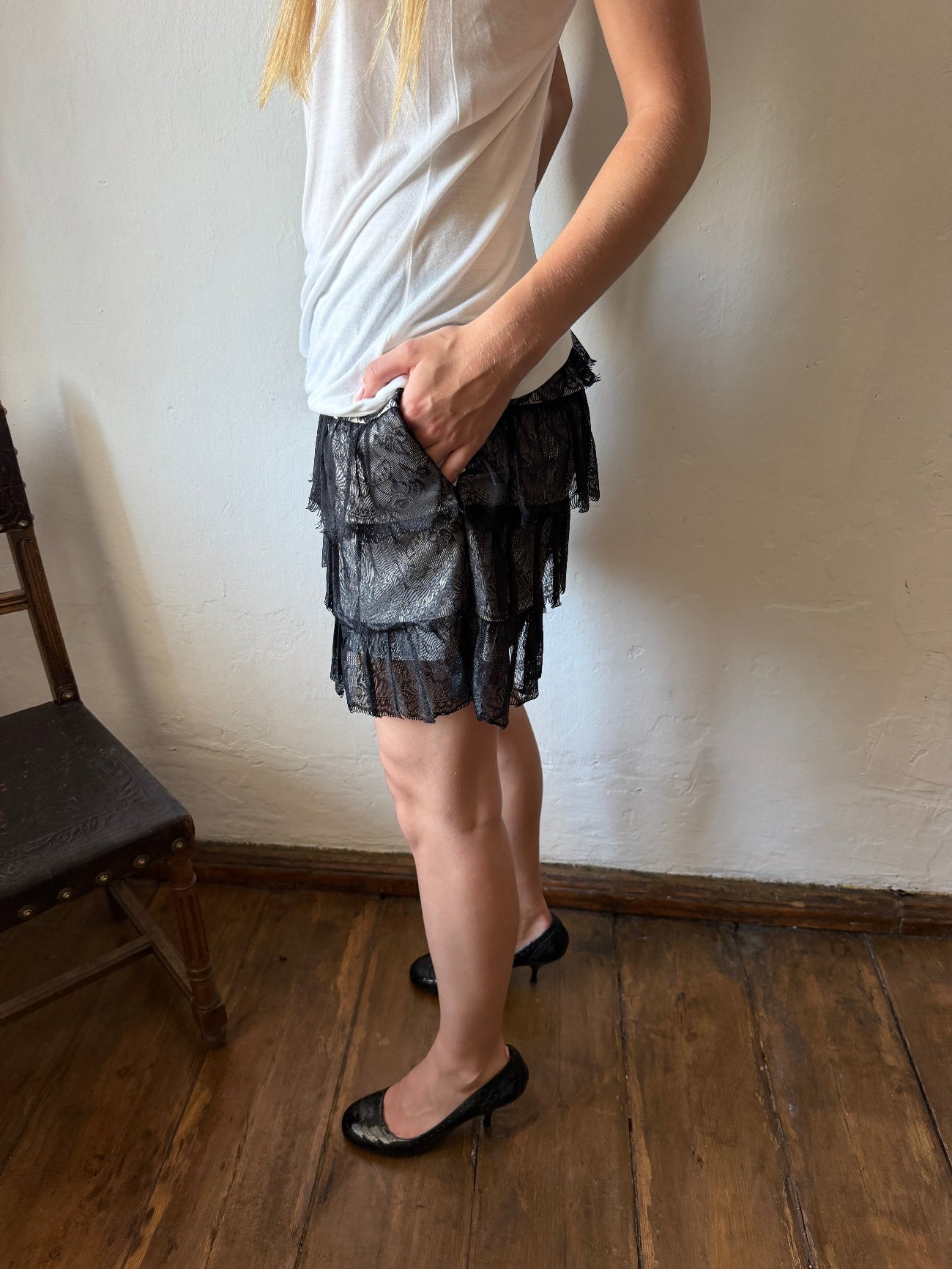 A person wearing a white sleeveless top and black tiered lace skirt, standing on a wooden floor against a plain wall.