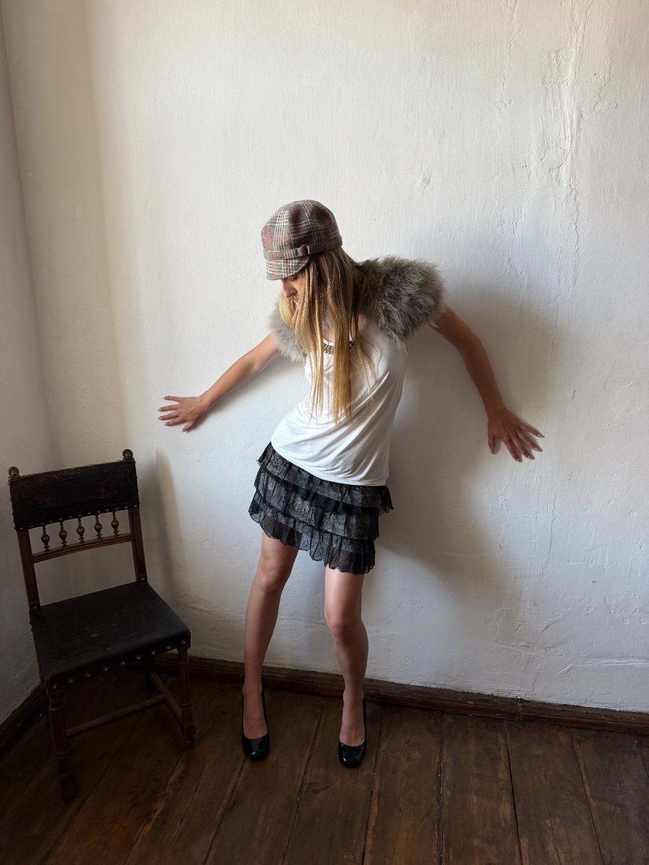 A woman posing in a room with a white wall and wooden floor, wearing a white t-shirt with a faux-fur collar, a dark patterned pleated mini skirt, a plaid hat, and black heels.