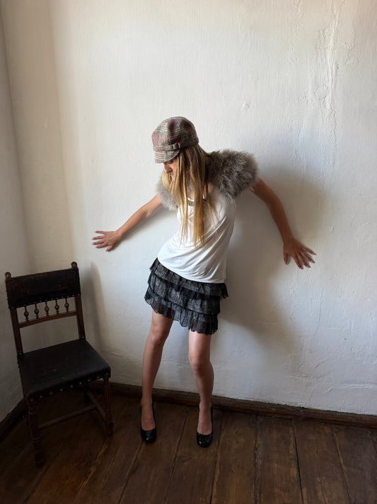 A woman posing in a room with a white wall and wooden floor, wearing a white t-shirt with a faux-fur collar, a dark patterned pleated mini skirt, a plaid hat, and black heels.