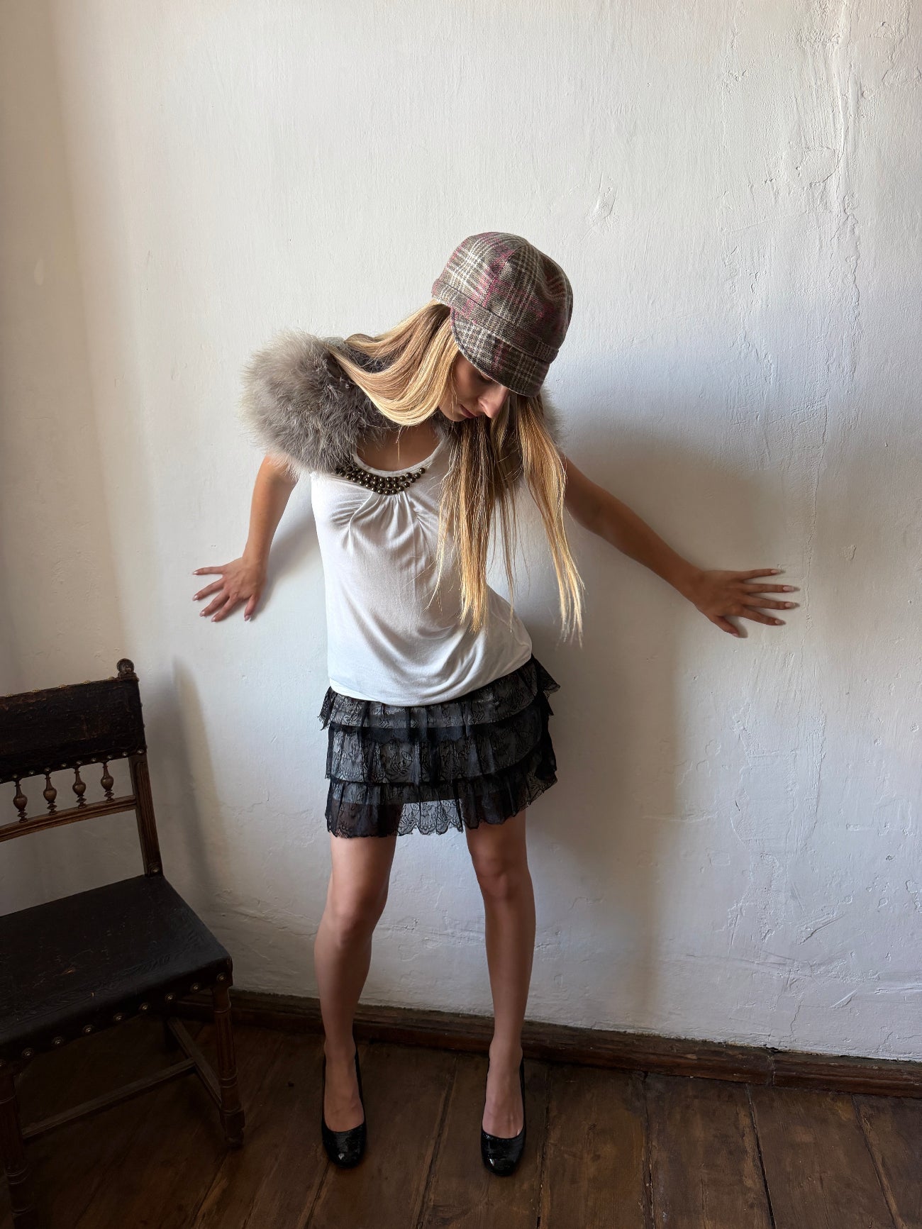 A woman posing in a white sleeveless top, a dark gray ruffled mini skirt, a plaid bucket hat, and a gray faux-fur collar against a white wall.